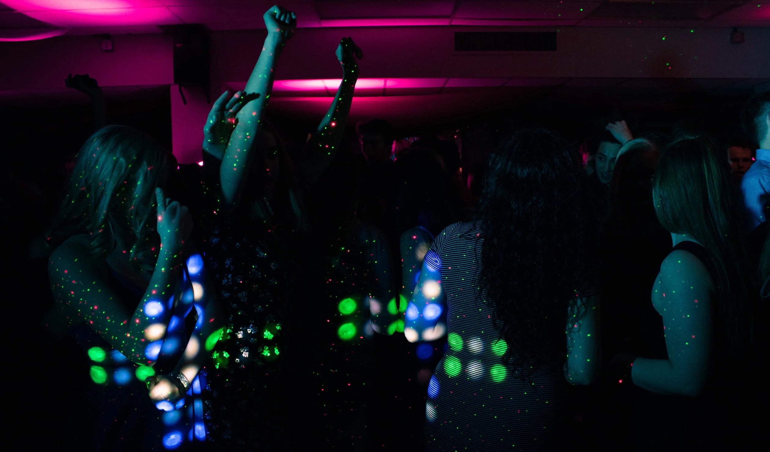 People dancing in a dark room with green, blue, and white laser lights and red LED lights in the background